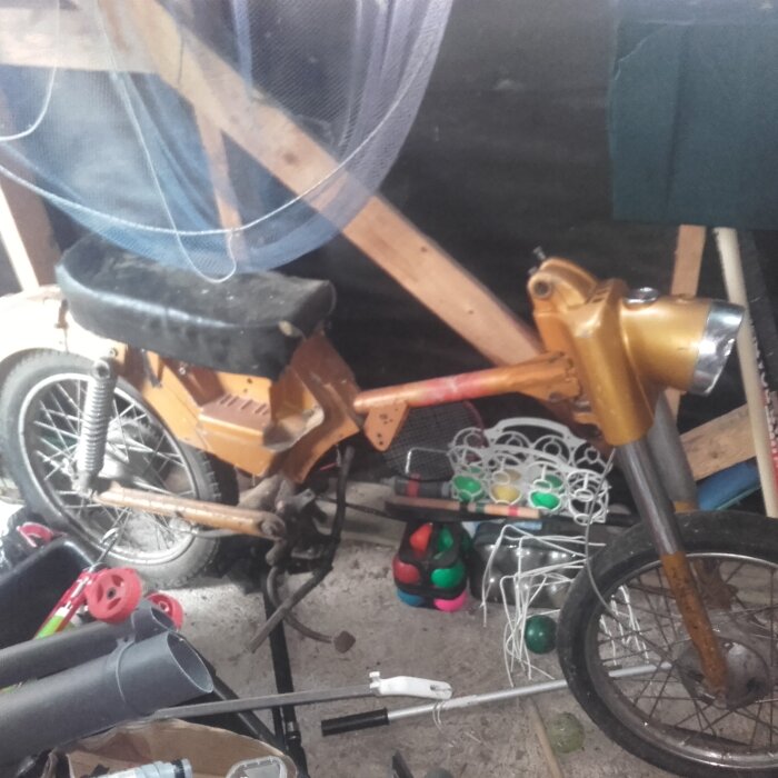 Gammal moppe i ett garage, rostiga och slitna delar, orange färg, omgiven av verktyg och annat bråte.