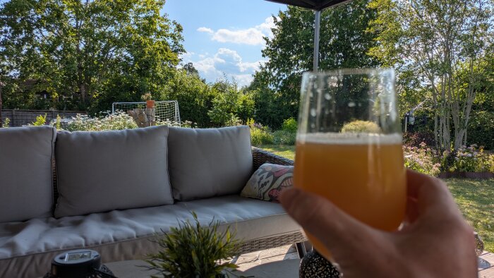 Hand håller ett glas Stigbergets El Capitano i skuggan under en pergola, med soffa och grönskande trädgård i bakgrunden.
