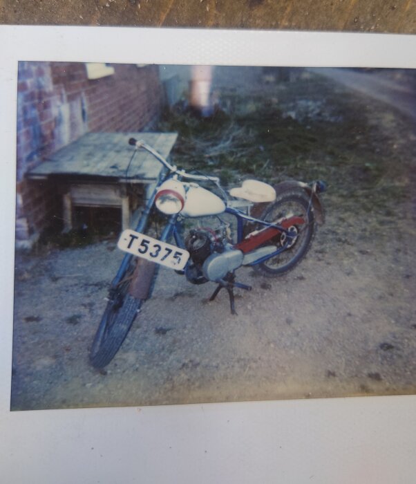 Motorcykel med registreringsskylt T5375, parkerad på grus bredvid byggnad.