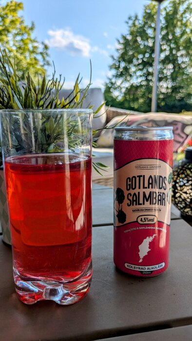Röd dryck i glas bredvid en burk med "Gotlands Salmbär" text, omgiven av grönskande bakgrund och klarblå himmel.