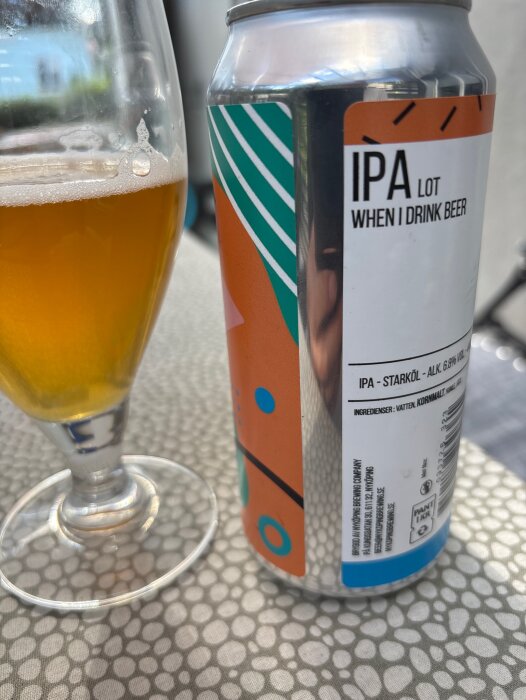 En ölburk med etikett "IPA lot when I drink beer" bredvid ett halvtomt glas med öl, stående på ett bord utomhus.