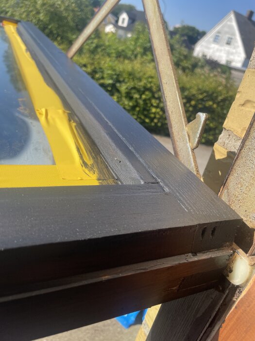 Närbild på ett svartmålat fönster med gul tejp längs kanten och en glaslist, utomhusmiljö med hus i bakgrunden.