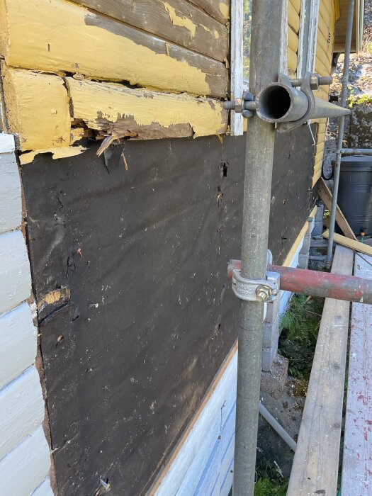 Fasadvägg med avskalad gul färg och rått trä synligt, delvis täckt av byggställning.