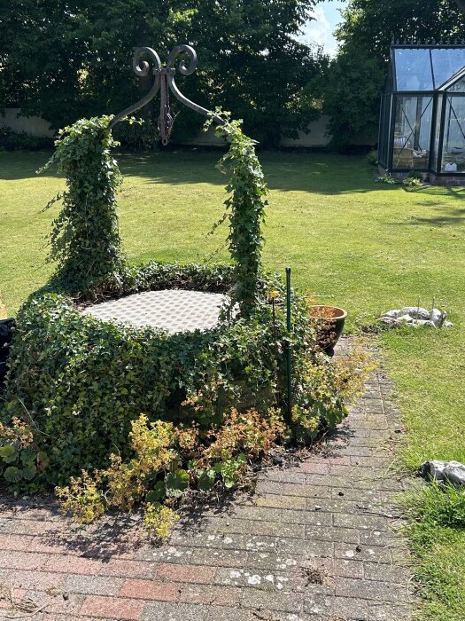 Gammal brunn täckt av murgröna och omgiven av stenlagda plattor i en grön trädgård med ett växthus i bakgrunden.