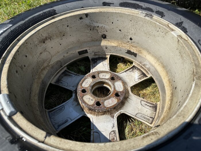 Smutsig Volvo-fälg före rengöring ligger på gräsunderlag, visar rost och smutsavlagringar på insidan av fälgen.