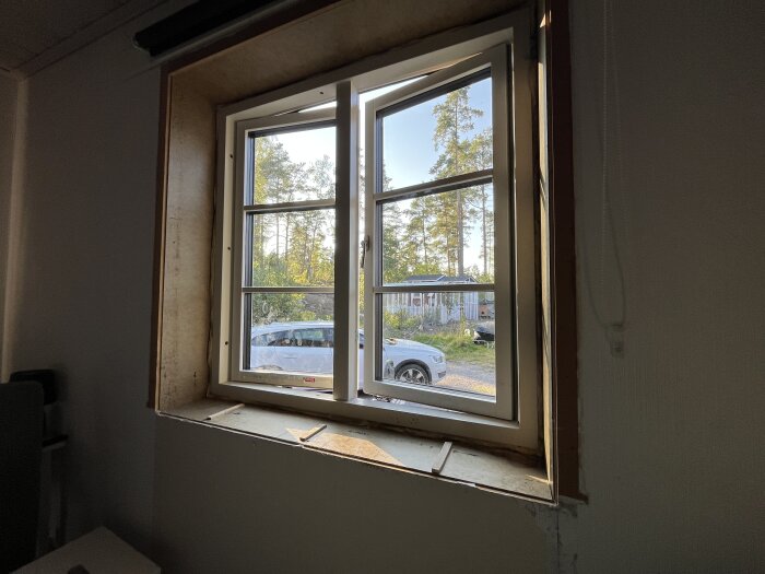 Öppet fönster med träkant inifrån, utsikt över gård med bil och träd i dagsljus.