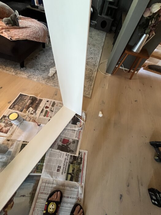 Målarprojekt i vardagsrum med fönsterram, tidningar för underlag, färgburk och pensel på golvet, samt en person i tofflor.