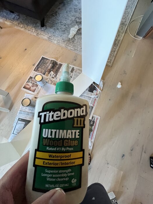 Titebond III trälimflaska hålls i handen, golv och målarprojekt synliga i bakgrunden tillsammans med målartidningar och burkar.