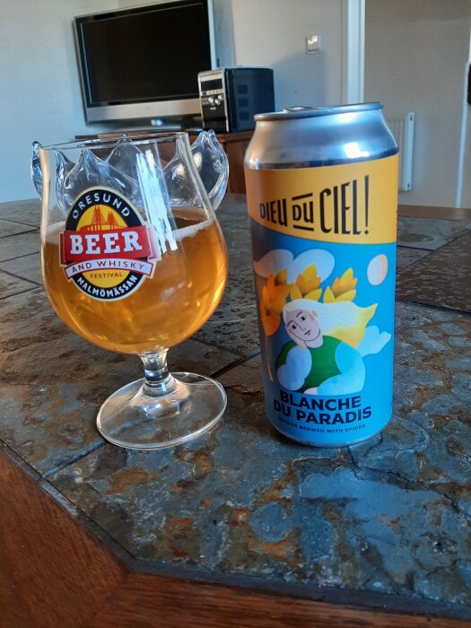 Ölglas med en gyllene dryck bredvid en burk med etiketten "Blanche du Paradis" och "Dieu du Ciel!", stående på ett bord inomhus.