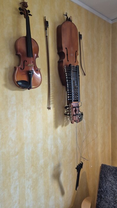 En nyckelharpa och en fiol hänger på väggen; nyckelharpans stränghållare har gått av, och strängarna hänger ner.