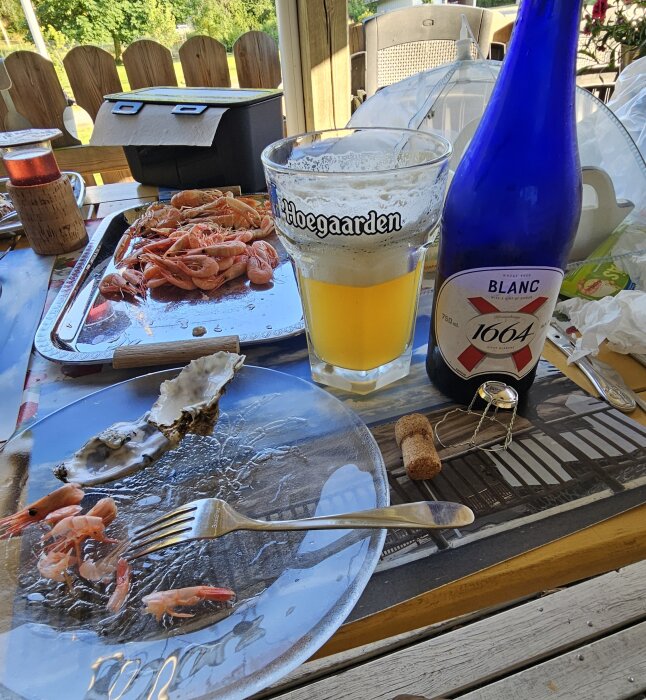 Flaska och glas med 1664 Blanc bier, tallrik med skaldjur och ostronskal på träbord utomhus.