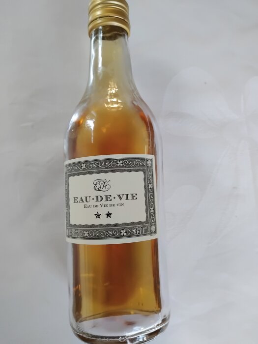 Flaska med eau de vie med etikett och guldlock på ett vitt underlag.