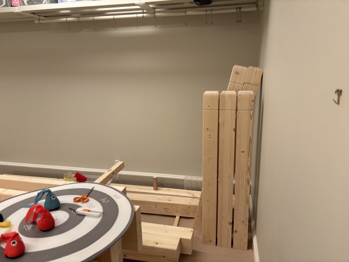 Högar av tillverkade trästolpar av limträ står lutade mot en vägg i ett rum med hobbyprojekt och små föremål på ett bord.