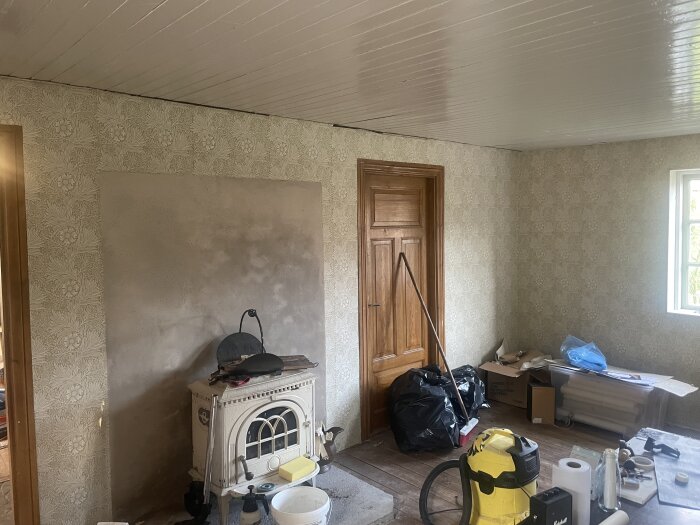 Rum med nymålad grå vägg intill en gammal kamin, omgiven av tapeter. Verktyg och städutrustning står på golvet.