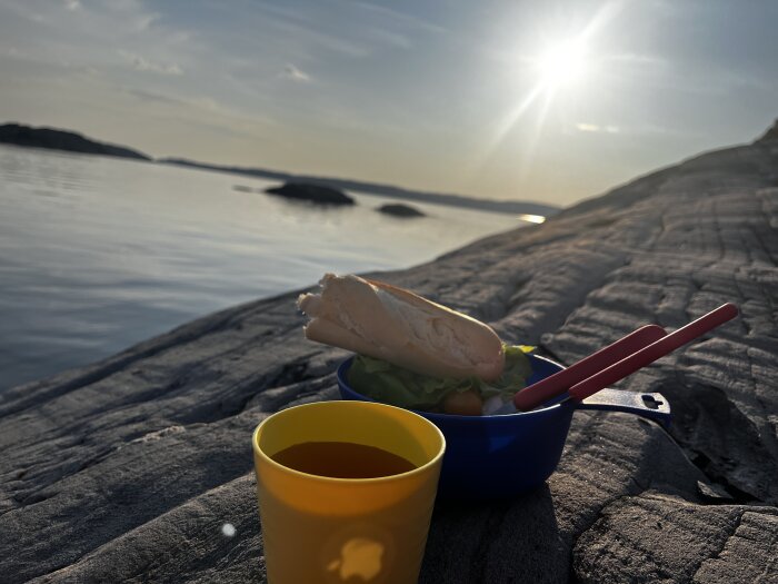 Picknickmat med baguette, sallad, och dryck på klippor vid solnedgång med havsutsikt.