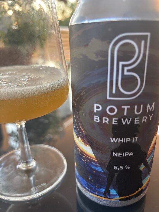 Glas med ljus öl och en burk med etikett "Potum Brewery Whip It NEIPA 6,5%" på ett bord.
