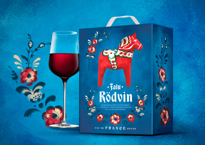 En vinbox med texten "Falu Rödvin" prydd med blomdesign och en dalahäst, tillsammans med ett glas rödvin mot en blå bakgrund.