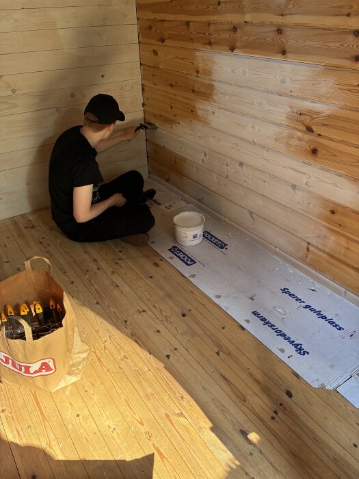 Person som målar en trävägg med vit färg i ett rum, omgiven av målarverktyg och skyddspapper på golvet.