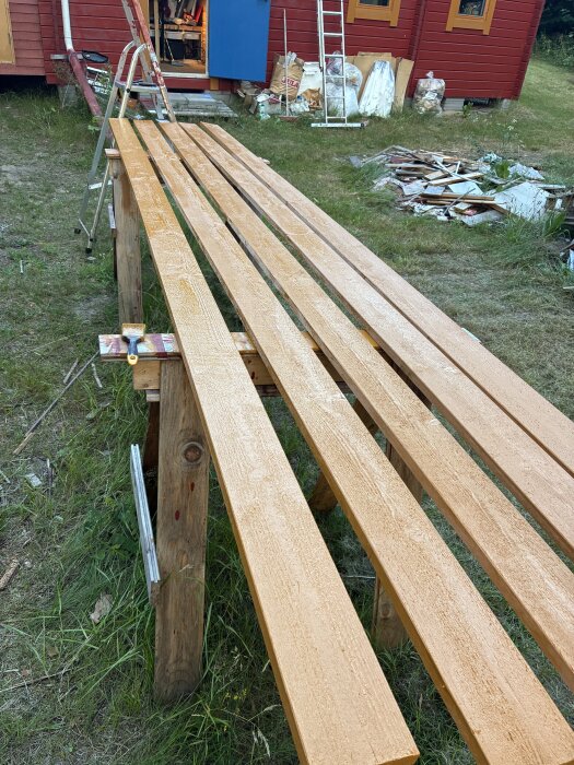 Brädor målade med gulockra linoljefärg ligger på bockar utomhus, med ett rött hus i bakgrunden.