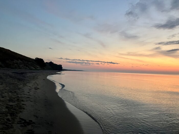 Strand i solnedgång med mjuka vågor som rullar in mot land. Himlen är färgad i pastellfärger och horisonten är svagt synlig.
