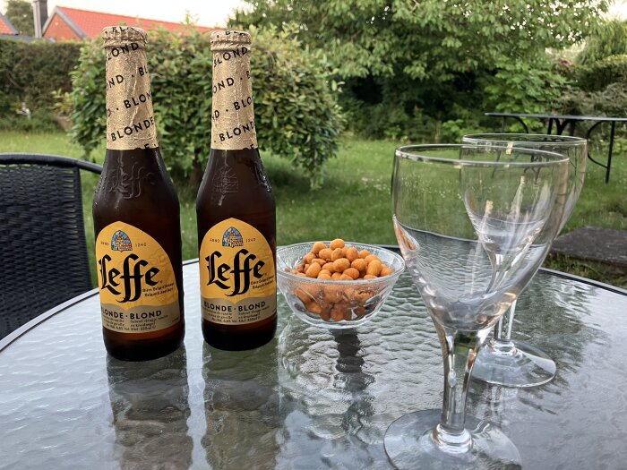 Två flaskor Leffe blonde öl, glas och snacks på ett trädgårdsbord.