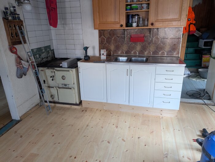 Nytt trägolv i kök med vedspis och vita köksskåp. Stege står bredvid spisen, och en borrmaskin samt hushållsartiklar är på bänken.