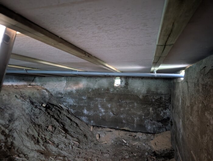 Källare med jordgolv, synliga rör i taket och en grundmur. Ljuset kommer in genom springor.