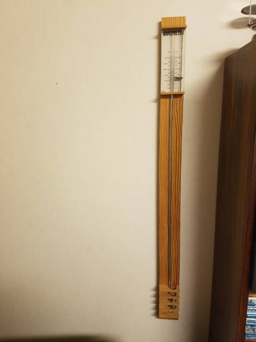 Kvicksilverbarometer monterad på en vägg, gjord av trä och med graderad skala upptill, mot en vit vägg bredvid en bokhylla.