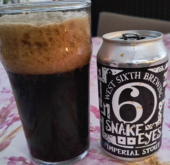 Glas med mörk Imperial stout och en ölburk från West Sixth Brewing Snake Eyes på ett blommigt underlag.