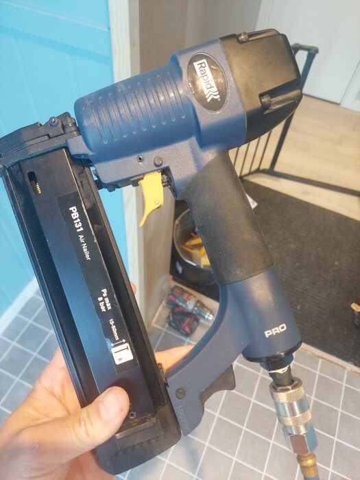 En blå dyckertpistol med svart handtag hålls i handen, på ett klinkergolv med verktyg i bakgrunden.