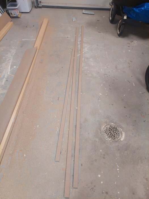 Flera smala träprofiler ligger på ett dammigt garagegolv efter sågning, tillsammans med verktygsspår och en golvbrunn i bakgrunden.