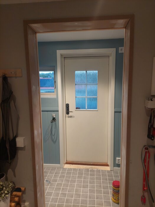 Vit dörr med glasruta och nytt dörrfoder i en hall, blå väggpanel och grått klinkergolv, städservetter står på golvet.