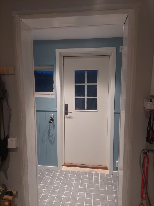 Entré med vitmålat dörr- och fönsterfoder, rektangulärt fönster i dörr och grått klinkergolv.