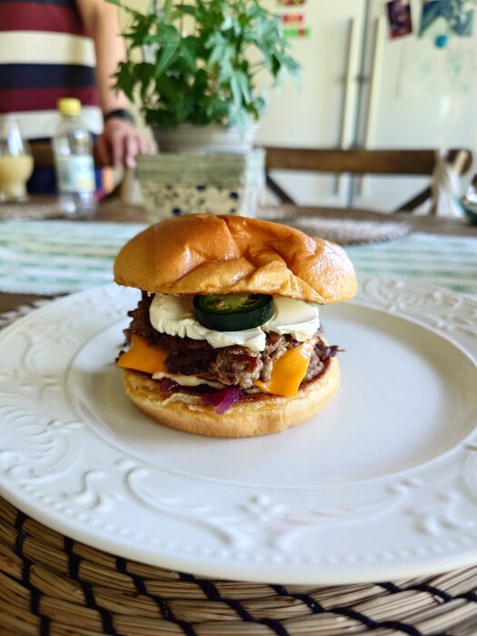 Hamburgare på vit tallrik, med köttfärs, smält ost, grön jalapeño och hemgjord dressing, i bakgrunden en växt och en person i randig tröja.