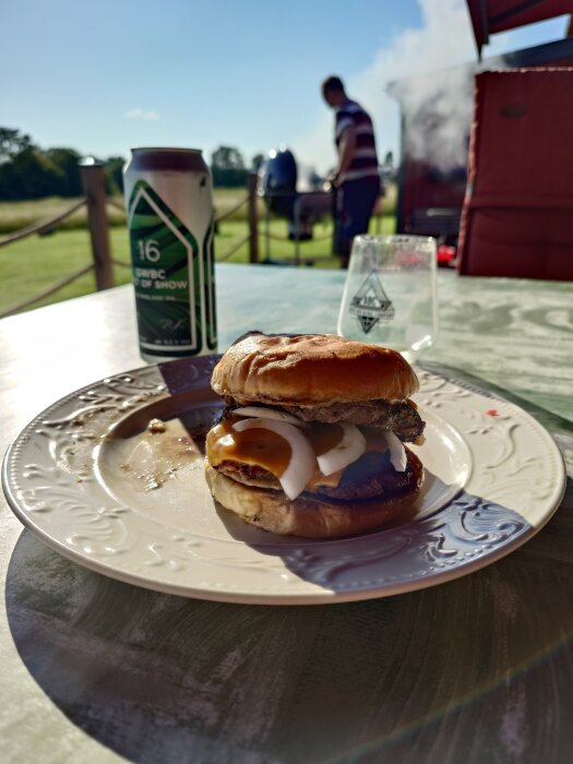 Hamburgare på tallrik med dressing och lök, ölburk bredvid på solbelyst uteplats, person vid grillen i bakgrunden.