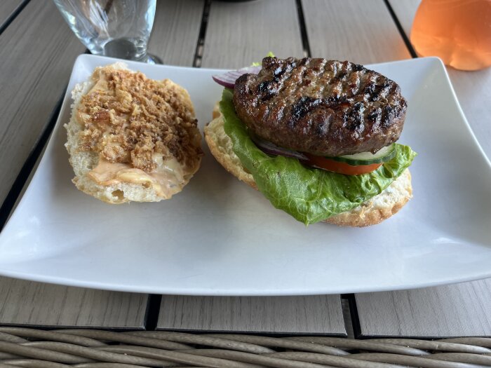 Grillad hamburgare med sallad, tomat, rödlök och dressing på en tallrik, delvis öppen med andra halvan täckt av rostad lök och dressing.