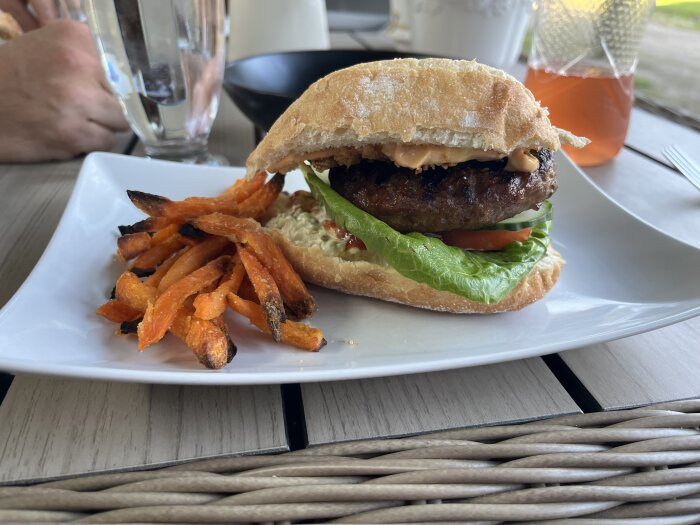 Grillad hamburgare med sallad, tomat, jalapenoost och dressing i bröd, serverad med sötpotatispommes på en vit tallrik.