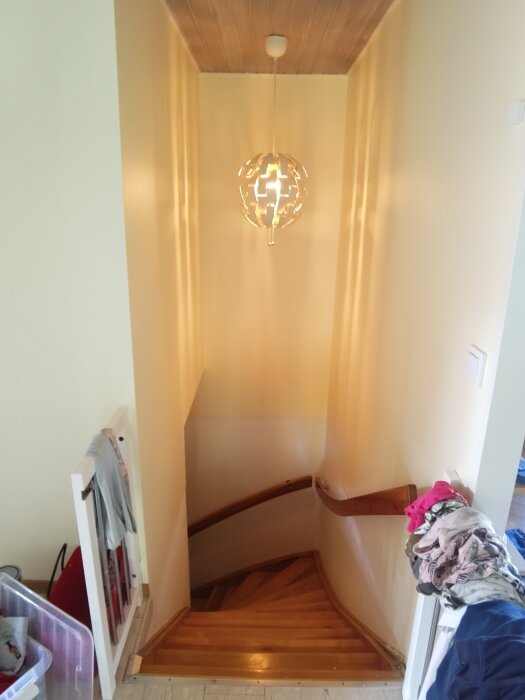 Trappa i källaren med ljus trätrappsteg och gulväggar, rund trälampa i taket och klädhängare intill trappräcke.