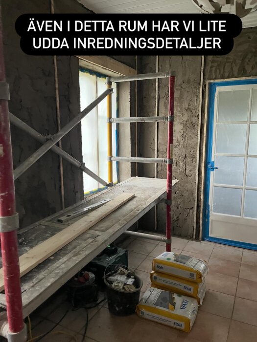 Renoveringsrum med byggställning, plankor och tre säckar byggmaterial på golvet. En dörr täckt av skyddsplast syns i bakgrunden.