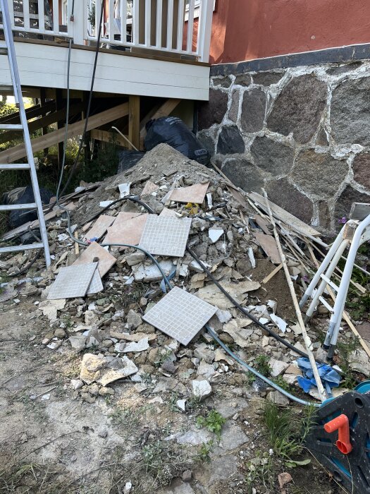 Hög med byggavfall utanför ett hus, bestående av betongbitar, klinkerplattor och träspill. En röd fasad och stentrappa syns i bakgrunden.