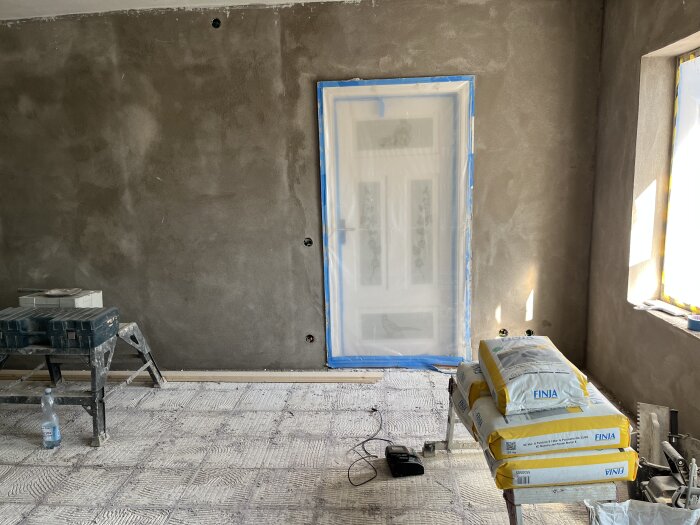 Rum under renovering med nyputsade väggar, täckt dörr, och byggmaterial på ett bord. Säckar märkt "Finja" syns intill ett fönster.