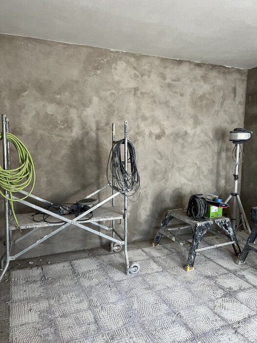 Rum under renovering med nyligen putsade och raka väggar, byggställning med kablar och verktyg, samt arbetslampa på stativ.