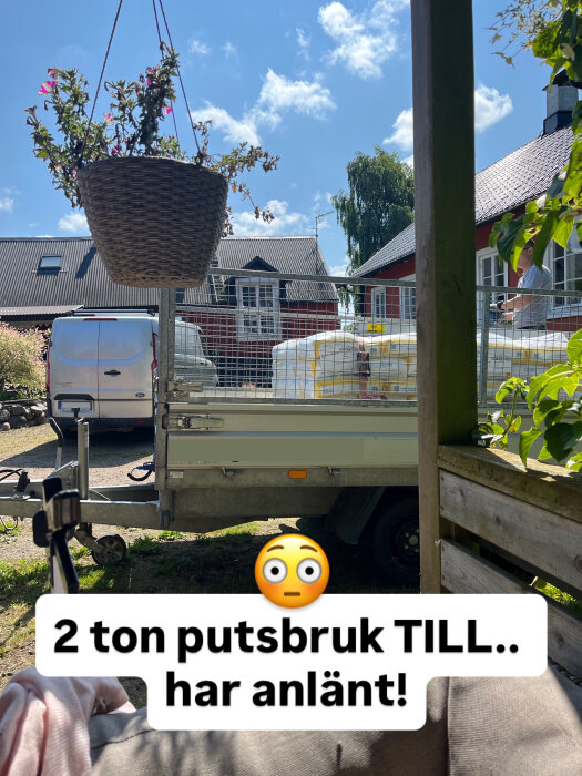 Släpvagn med säckar av putsbruk och en hängande blomkruka utanför hus. Text: "2 ton putsbruk TILL.. har anlänt!".