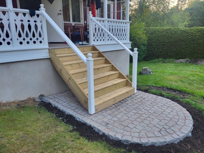 Nybyggd trätrappa med vita handledare och stolpar, lagd mot husets ingång med plattlagd stenyta framför, omgiven av gräsmatta och buskage.
