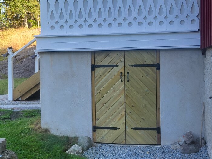 Träfärgade dubbeldörrar med svarta beslag leder till källaren under en vit dekorerad veranda, gräsmatta och trappa syns delvis.