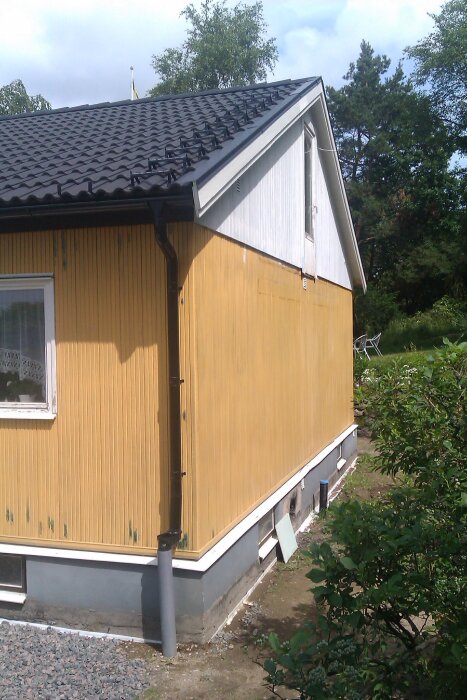 Gulmålad husfasad med synliga korrosionsbubblor, svart tak, och vit gavel. Omgiven av grönska och grusbelagd grund.