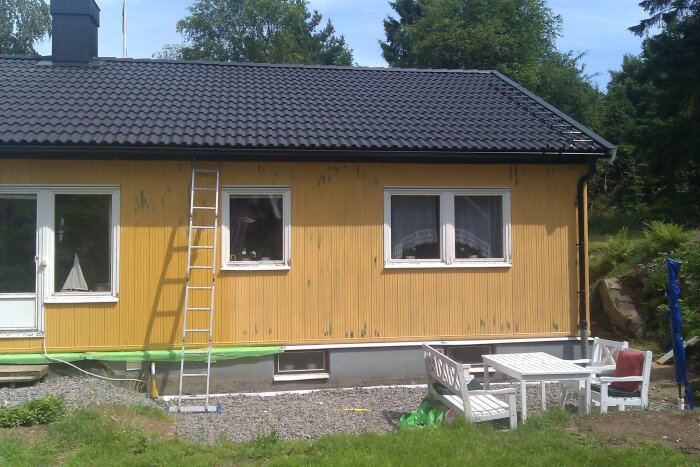 Gult hus med köksentré, stege, plastförpackad sockel och vit trädgårdsmöbel. Fasad visar korrosionsfärg och bubblor från renovering.