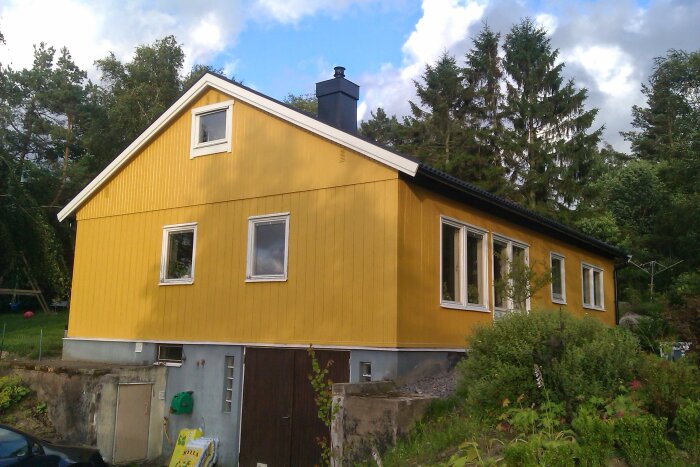 Gult hus från 2025 med grönska i förgrunden, visar aluminiumfasad och flera fönster under blå himmel med moln.