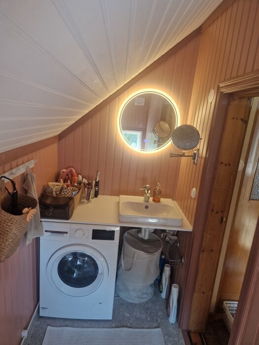 Badrum med tvättmaskin under en bänk, handfat ovanpå, och runda spegeln mot snedtaksvägg; golvet är i klinker.