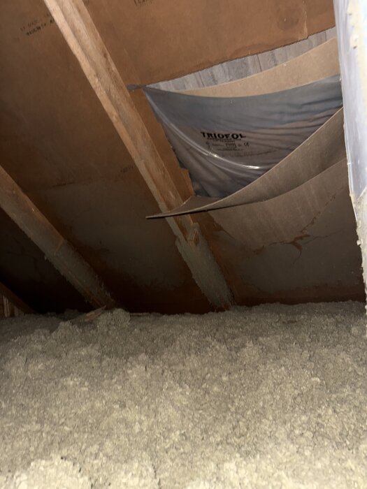Knäckt masonit på vinden med rivet folie avtäckt, visar isolering och undersida av betongpannor.
